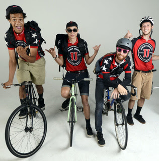 Four Jimmy John’s delivery cyclists in uniform, each with a bicycle and a delivery bag, pose for a studio photo. They wear black and red cycling jerseys with the Jimmy John’s logo, helmets, and sunglasses. The mood is energetic and fun.