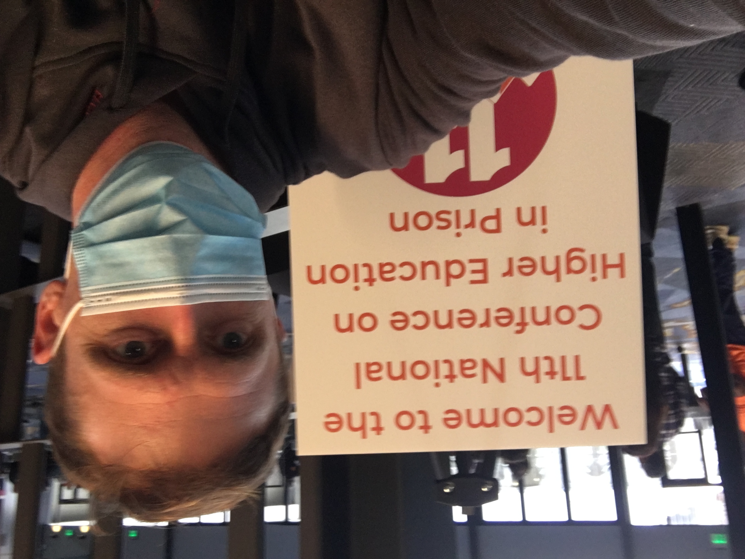 John Wise, wearing a medical mask and a hoodie, takes a selfie in front of a sign that reads “Welcome to the 11th National Conference on Higher Education in Prison.” The setting appears to be a conference venue, with people in the background.