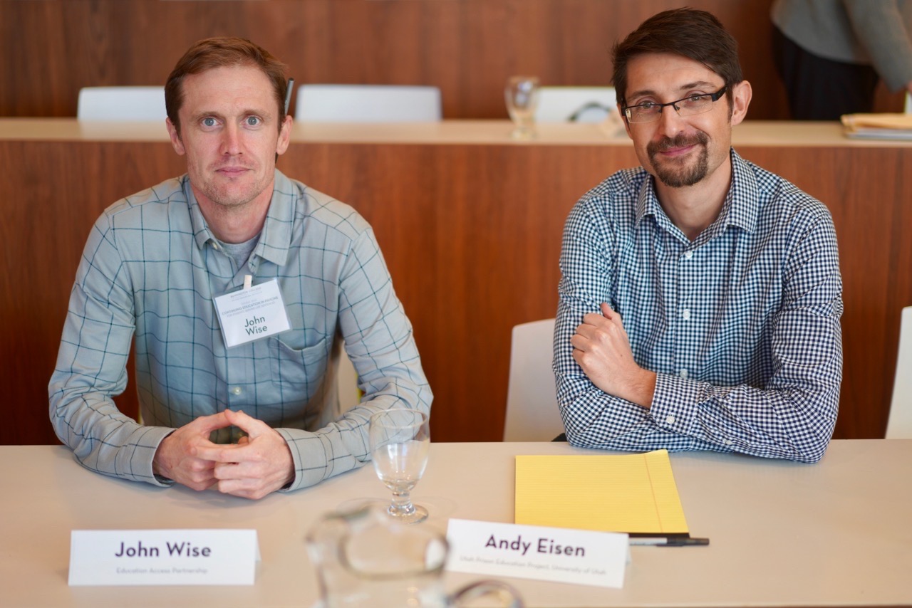 John Wise and Andy Eisen sit side-by-side at a conference table. Both have name placards in front of them (John Wise, Education Access Partnership; Andy Eisen, University of Utah Prison Education Project). They are indoors, in a warmly-lit meeting room with wood paneling.