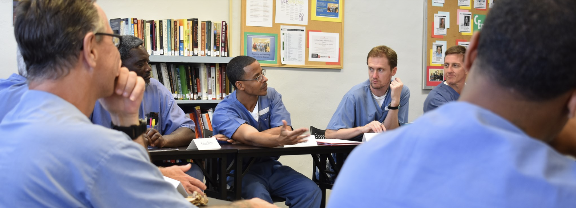 John Wise seated and engaged in a group discussion in a classroom as part of the Community Education Project at Tomoka Correctional Institution. Other participants in blue uniforms are visible, with bookshelves and bulletin boards in the background.