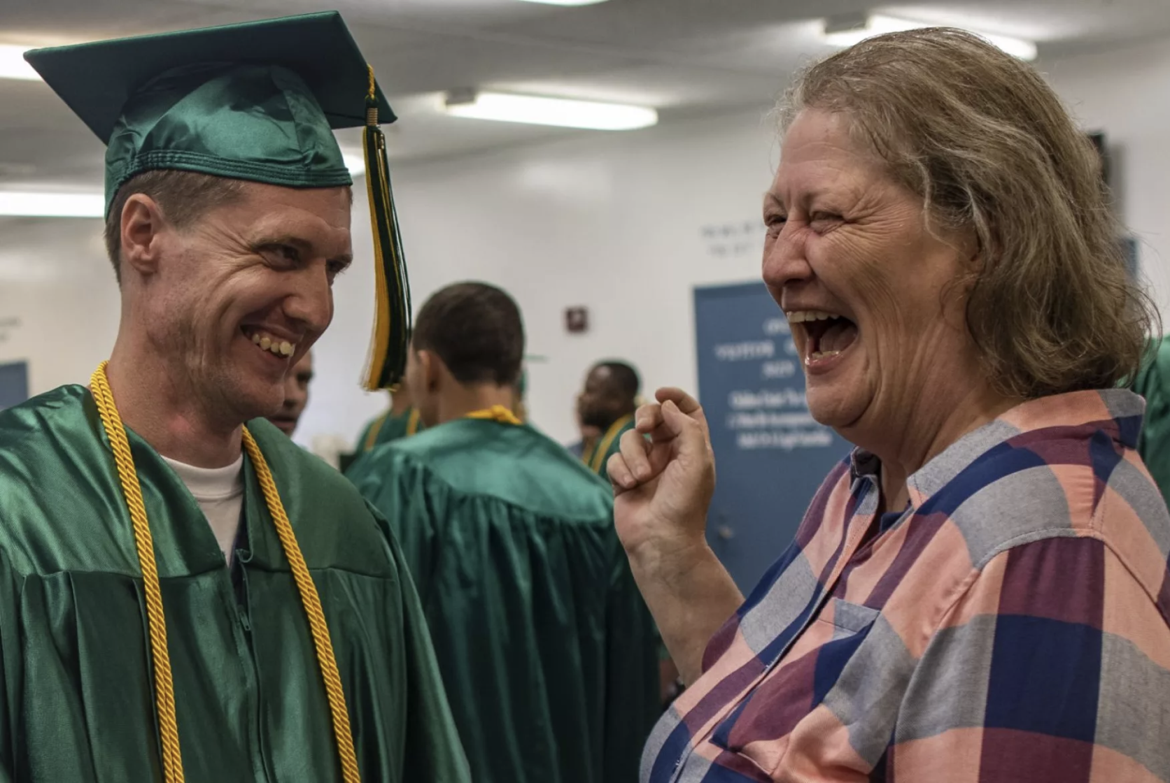 John Wise, wearing a green cap and gown with gold honor cords, shares a joyful, laughing moment with his mother at graduation. Both are beaming; his mother is dressed in a plaid shirt and her expression is one of pride and happiness.