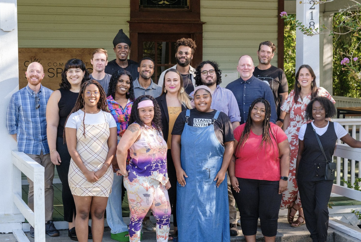 A group portrait on a porch with diverse participants of Community Spring, including John Wise, standing among other fellows and staff outside a house. The group is smiling and appears relaxed and happy, gathered in daylight in front of a “Community Spring” sign.