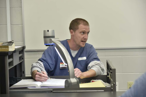 John Wise sits at a classroom desk, mid-sentence, holding a pen and a textbook, with a document camera arm in front of him. He is dressed in a blue uniform, teaching a lesson to others not visible in the photo.